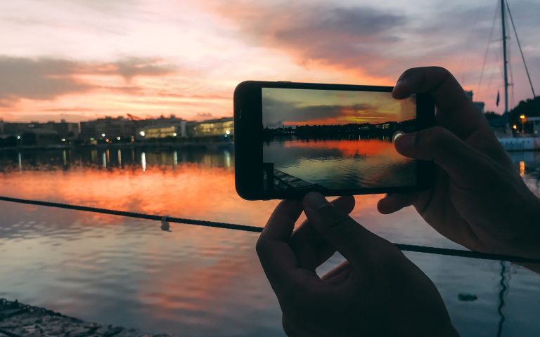 Man hands taking a picture with mobile phone of a beautiful sunset in a harbor